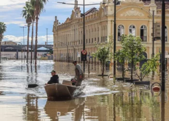 Homem e mulher em um barco enfrentando a enchente, ilustrando os impactos das chuvas e a necessidade de auxílio para reconstrução.