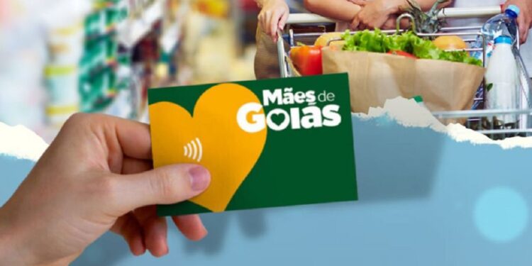 Cartão do programa Mães de Goiás sendo segurado por uma mão, com logo visível e fundo de compras.