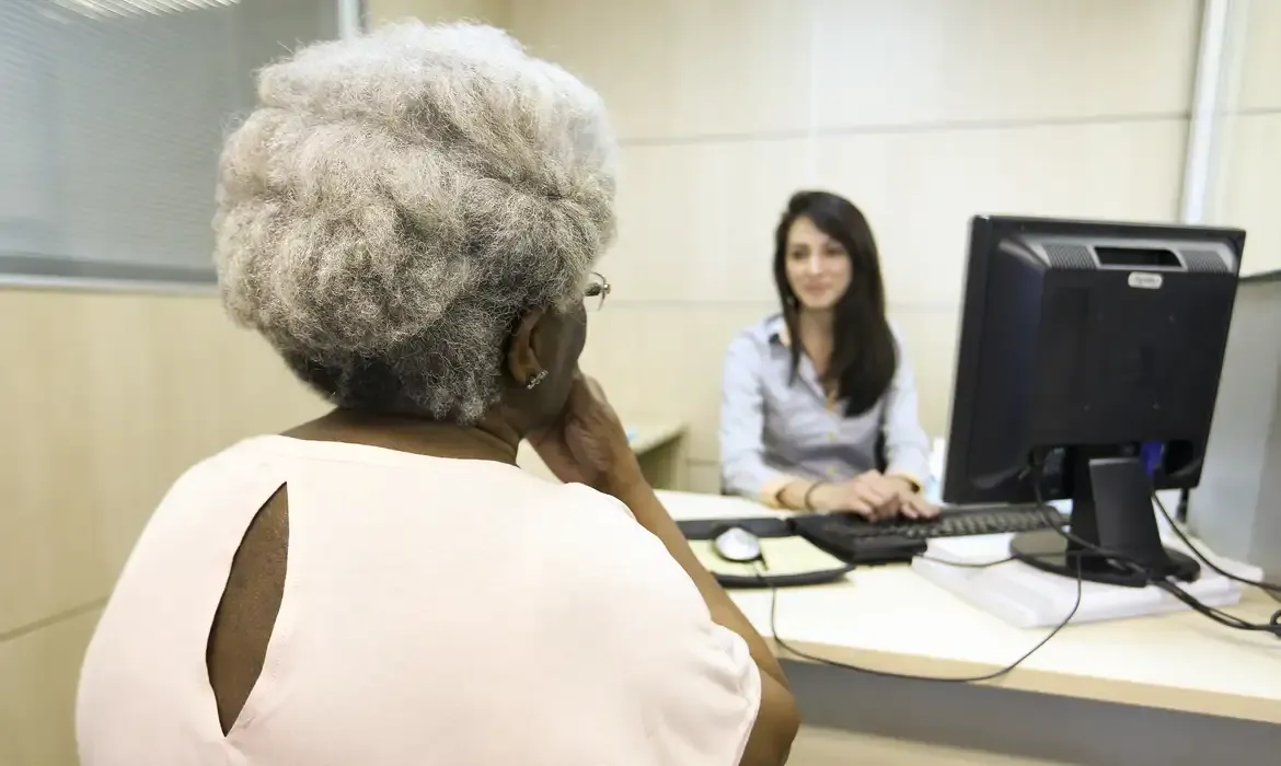 Idosa sendo atendida por assistente em escritório, representando serviços de assistência social.