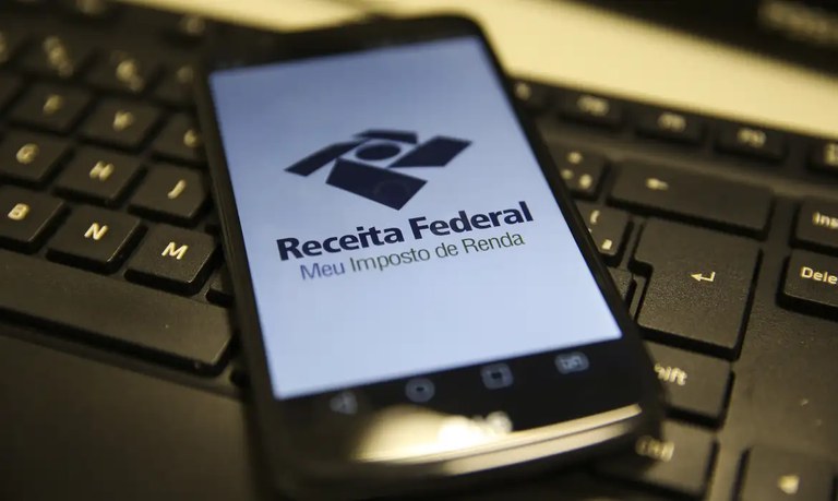 Imagem de um celular exibindo o logo da Receita Federal e a inscrição 'Meu Imposto de Renda' sobre um teclado.