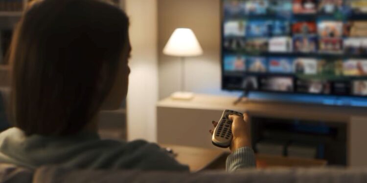 Mulher assistindo TV em casa, com controle remoto, simbolizando a transição do sinal analógico para o digital.
