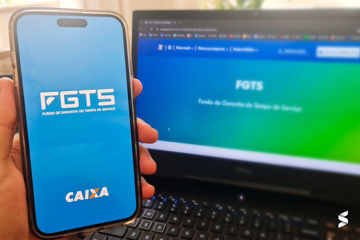 Celular com tela do aplicativo FGTS da Caixa, ao lado de laptop com página da Caixa aberta
