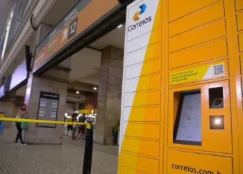 Totem eletrônico dos Correios instalado em uma agência com pessoas ao fundo.