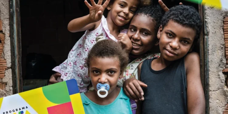 Família beneficiária do Bolsa Família sorrindo, com cartão do programa visível, em ambiente simples.