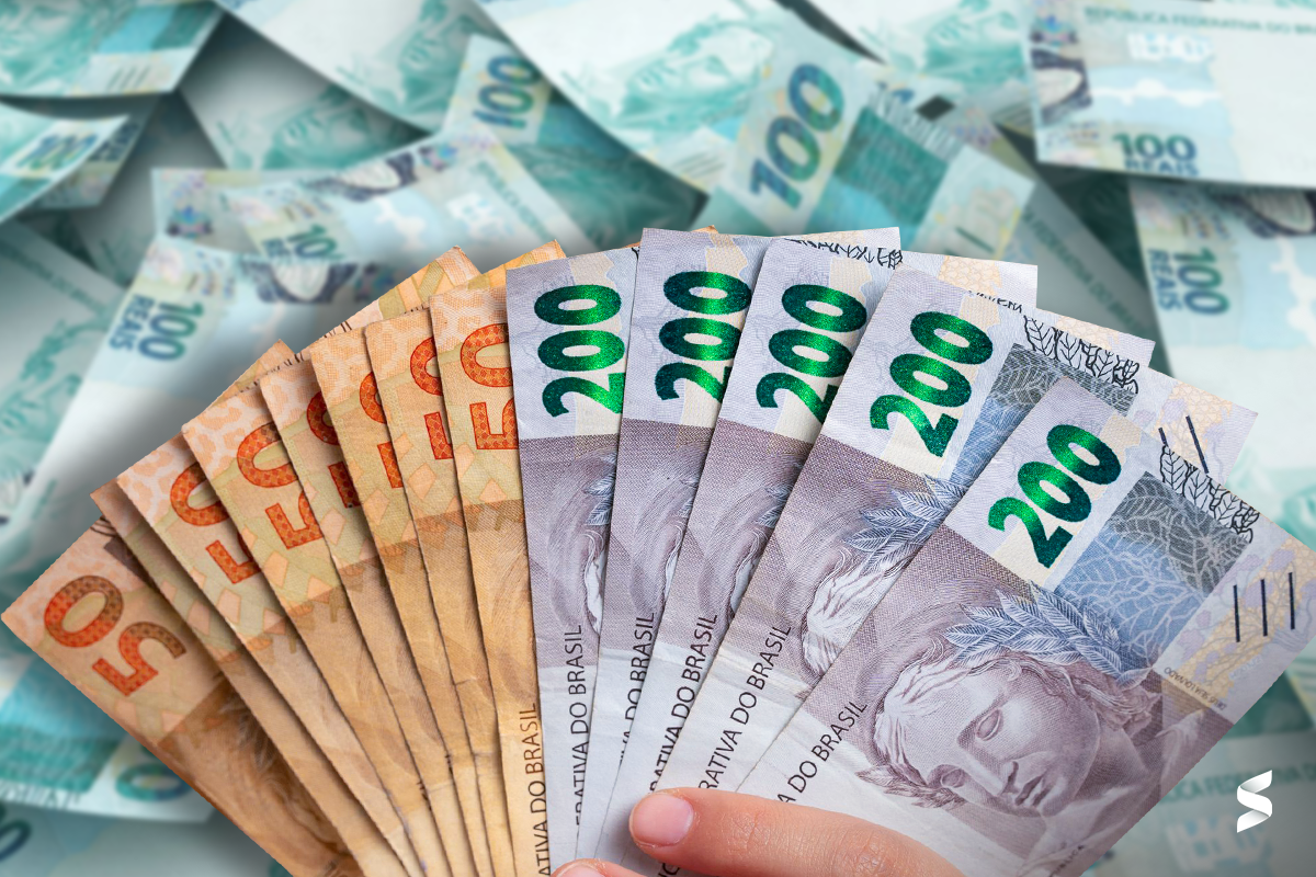 Mão segurando cédulas brasileiras de 50 e 200 reais com fundo desfocado de notas de 100 reais.