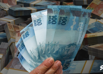 Mão segurando várias cédulas de 100 reais com muitas pilhas de dinheiro ao fundo.