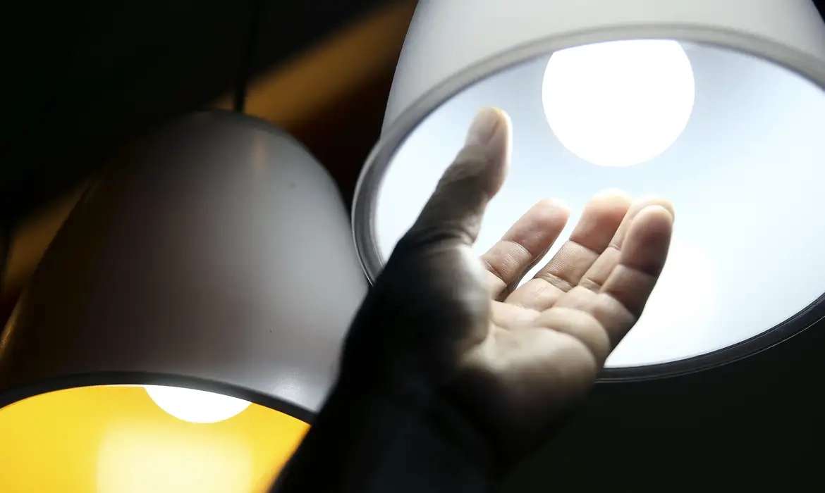 Mão estendida próxima a uma lâmpada acesa, simbolizando o aumento do custo de energia.