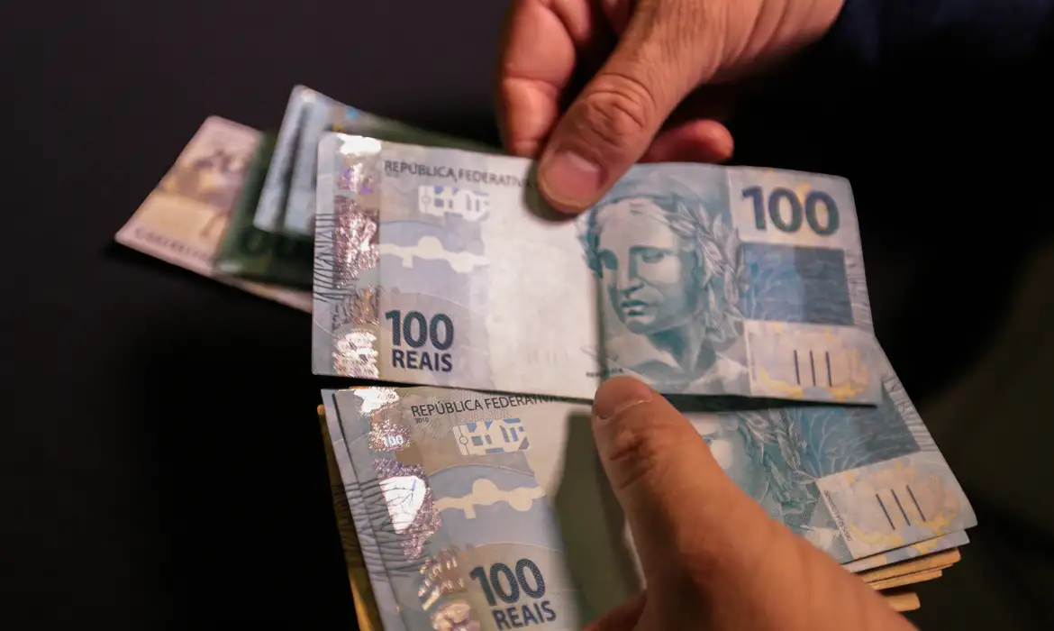 Mãos contando cédulas de 100 reais, ilustrando o aumento da multa por atraso no Imposto de Renda