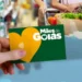 Cartão do programa Mães de Goiás sendo segurado por uma mão, com fundo de compras no supermercado.