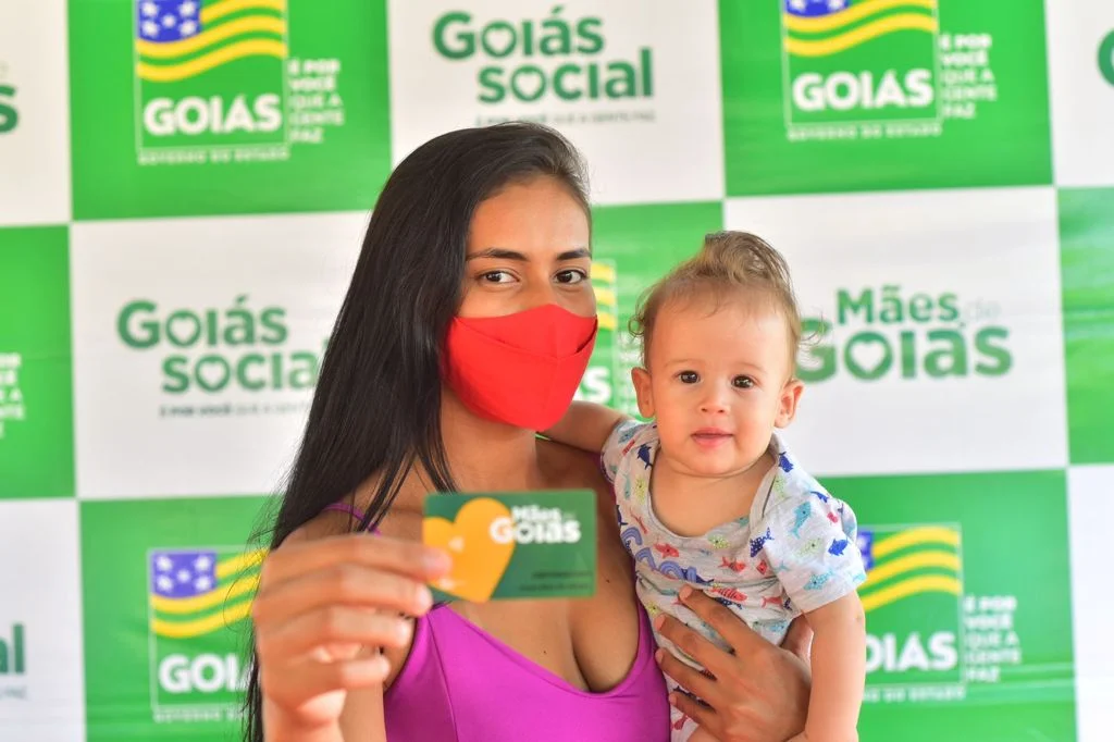 Mulher segurando bebê com cartão do Programa Mães de Goiás, em frente a um fundo com logotipos do programa e do Governo de Goiás.