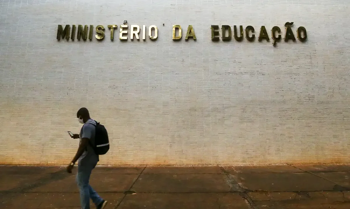 Homem caminhando em frente ao Ministério da Educação enquanto consulta o celular