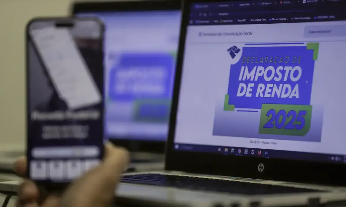 Tela de computador exibindo o site da Declaração de Imposto de Renda 2025, com uma pessoa segurando um celular em segundo plano.