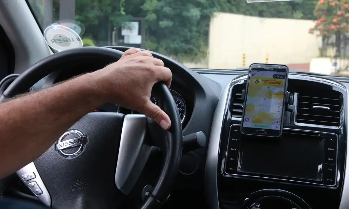 Motorista de aplicativo com o celular no suporte do carro, mostrando um mapa e indicando uso de aplicativo de navegação.
