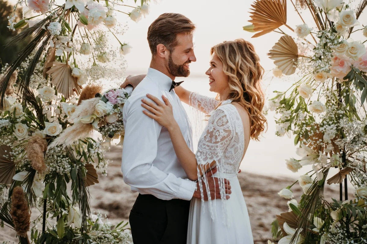 Casamento na praia com casal sorrindo e flores ao fundo