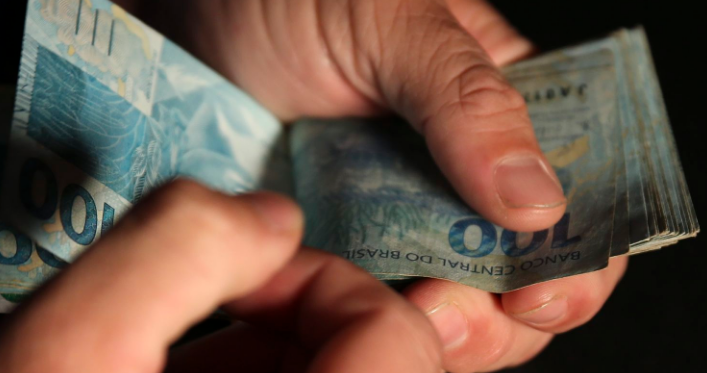 Mãos contando notas de 100 reais, ilustrando o impacto do novo salário mínimo de R$ 2,2 mil.