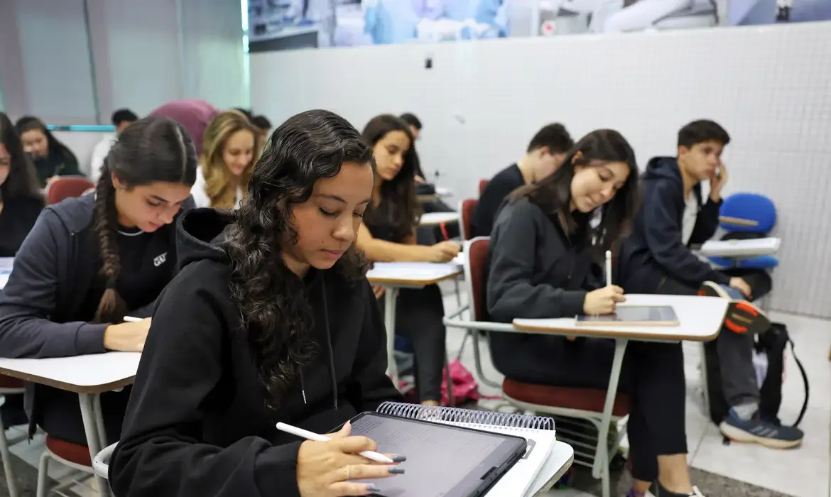 Alunos de diferentes idades estudando em sala de aula com tablets e materiais de estudo.