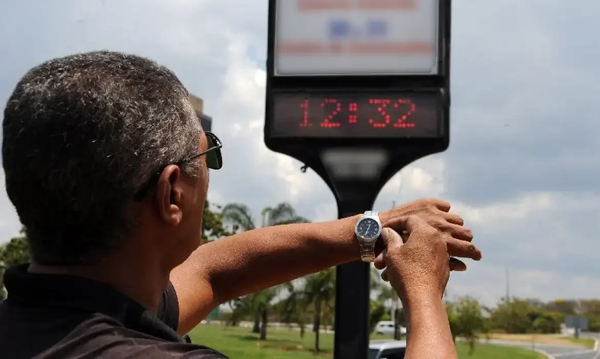 Pessoa verificando o horário de verão em um painel eletrônico e no relógio pessoal.