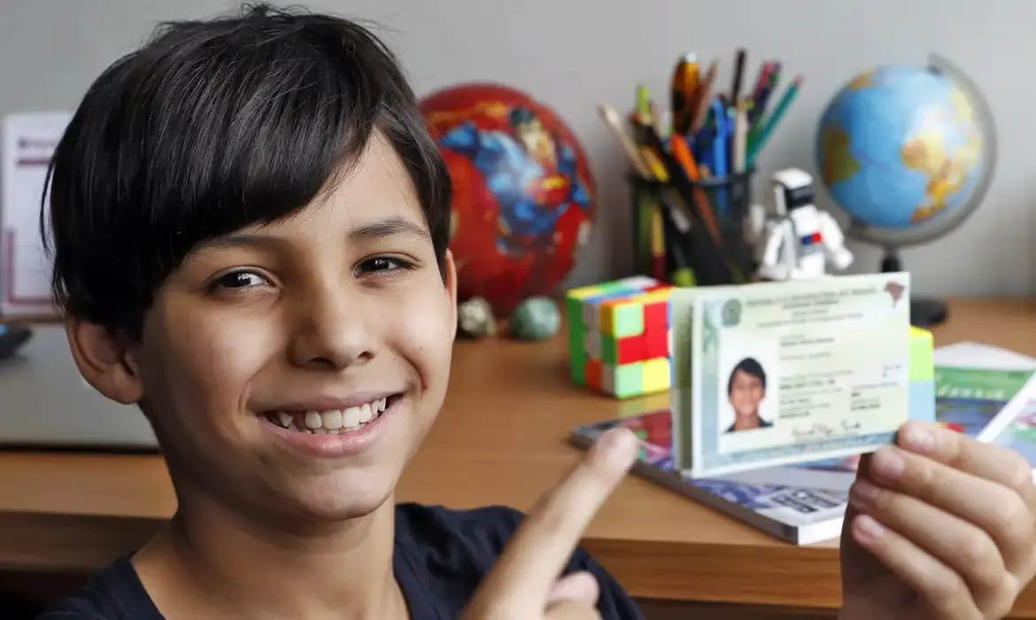 Criança segurando documento de identidade com um sorriso, em um ambiente de estudo com elementos de educação ao fundo.