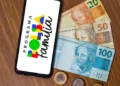 Celular exibindo a marca do Programa Bolsa Família sobre mesa de madeira, ao lado de cédulas de 100, 50 e 20 reais e moedas, em referência ao calendário de pagamentos de agosto.