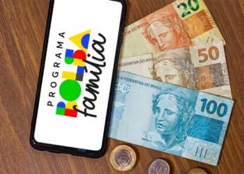 Celular exibindo a marca do Programa Bolsa Família sobre mesa de madeira, ao lado de cédulas de 100, 50 e 20 reais e moedas, em referência ao calendário de pagamentos de agosto.
