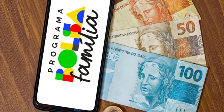 Celular exibindo a marca do Programa Bolsa Família sobre mesa de madeira, ao lado de cédulas de 100, 50 e 20 reais e moedas, em referência ao calendário de pagamentos de agosto.