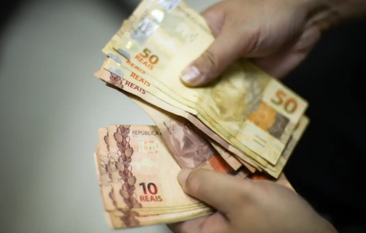 Mãos trocando notas de 10 e 50 reais com fundo desfocado, representando pagamento do Abono Salarial PIS 2025.