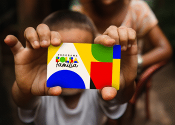 Mãos de uma criança segurando o cartão do programa Bolsa Família, com o novo design colorido e a logo do governo. A imagem representa o apoio financeiro e a inclusão social oferecidos a milhões de famílias no Brasil.