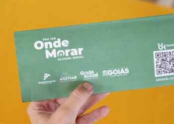 Pessoa segurando envelope verde do programa Aluguel Social de Goiás, auxilio aluguel com QR Code e logos de apoio.