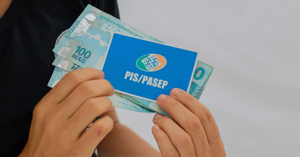 Pessoa segurando o cartão PIS/PASEP junto a cédulas de R$ 100, simbolizando o direito ao abono salarial de 2025.