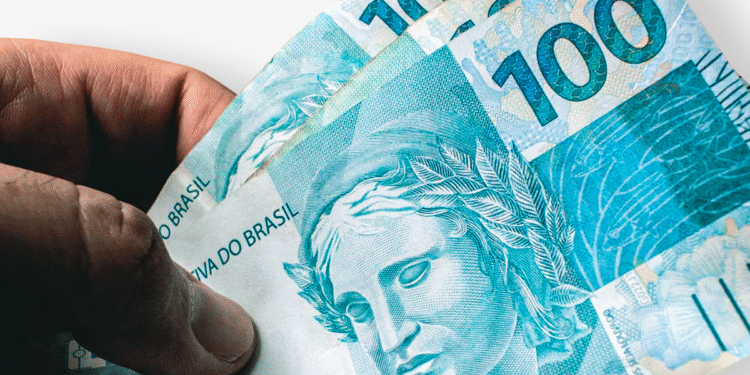 Mão segurando três notas de 100 reais, representando o auxílio de R$ 2 mil do governo.