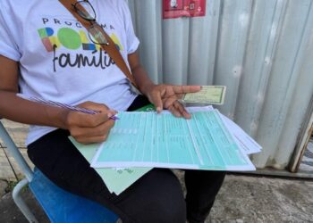 Visita domiciliar preenchendo formulário relacionado ao Bolsa Família, sentada em uma cadeira externa.