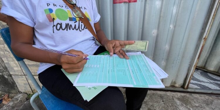 Visita domiciliar preenchendo formulário relacionado ao Bolsa Família, sentada em uma cadeira externa.