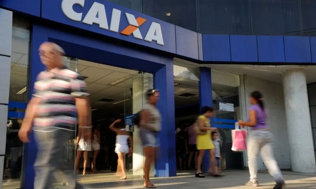Pessoas caminhando em frente a uma agência da Caixa Econômica, demonstrando movimento e acessibilidade.