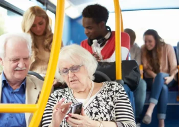 Imagem de idosos em um transporte público, com foco na tela do celular de uma idosa que interage com a interface, enquanto outros passageiros estão ao fundo.