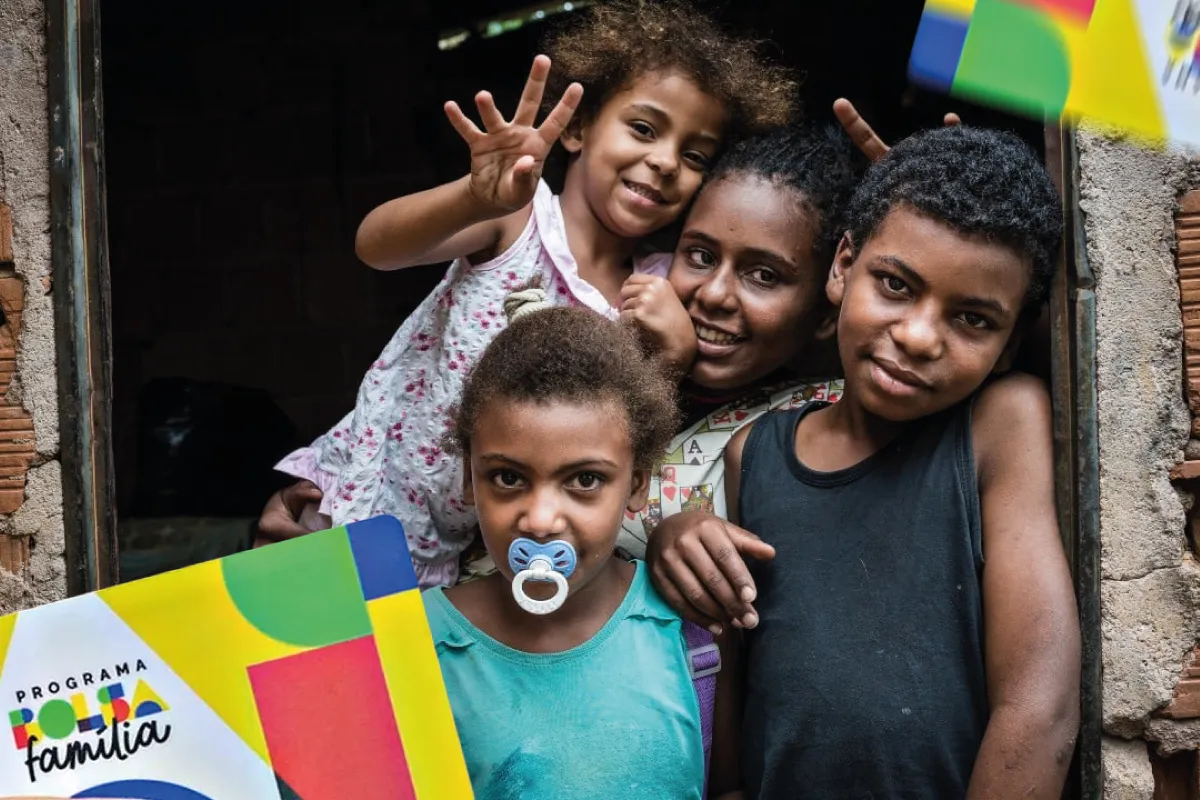 Crianças e jovens sorrindo em frente a uma casa, com a imagem de um cartão do Programa Bolsa Família na parte inferior esquerda da foto.