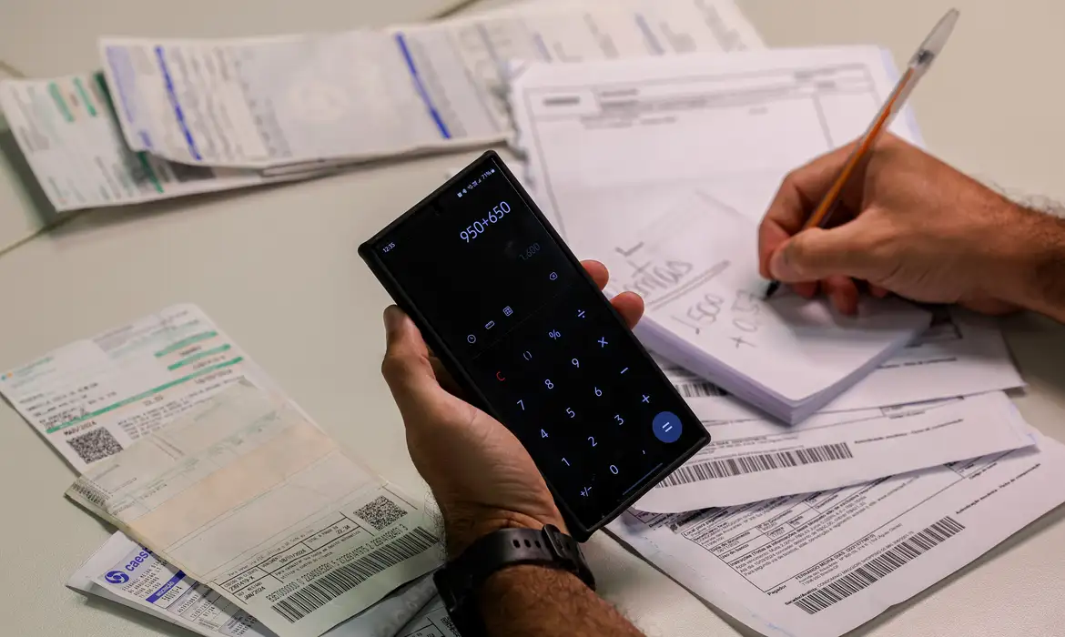 Pessoa calculando contas com celular e boletos sobre a mesa