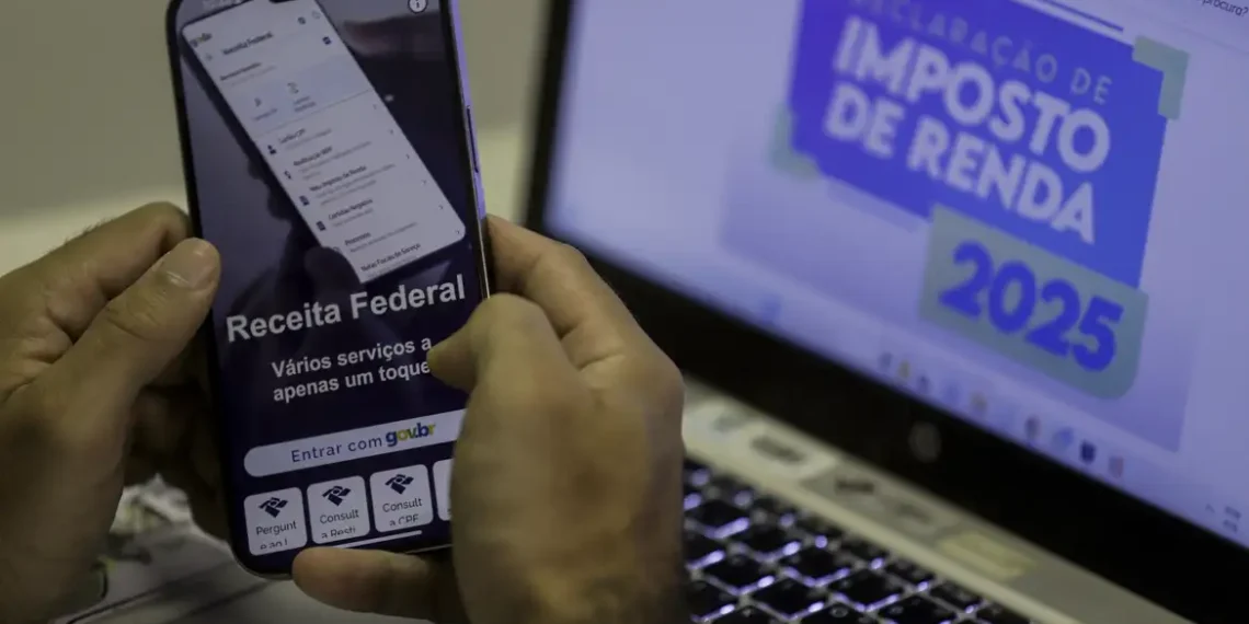 Aplicativo da Receita Federal em smartphone com computador exibindo "Declaração de Imposto de Renda 2025" ao fundo.