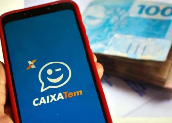 Aplicativo CAIXA Tem na tela de smartphone, com notas de R$ 100 reais ao fundo. Imagem simboliza os pagamentos do Bolsa Família e Auxílio Gás.