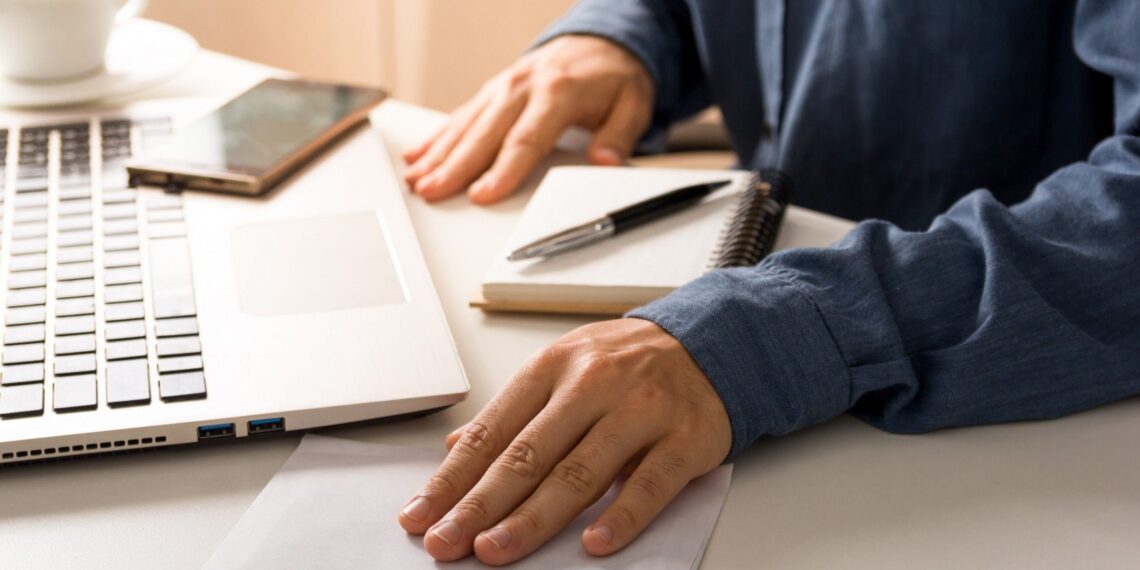 Imagem de uma mesa de trabalho com um computador portátil, um celular, uma xícara e uma folha com dinheiro de R$ 100 visível, representando uma atividade financeira ou transação. 