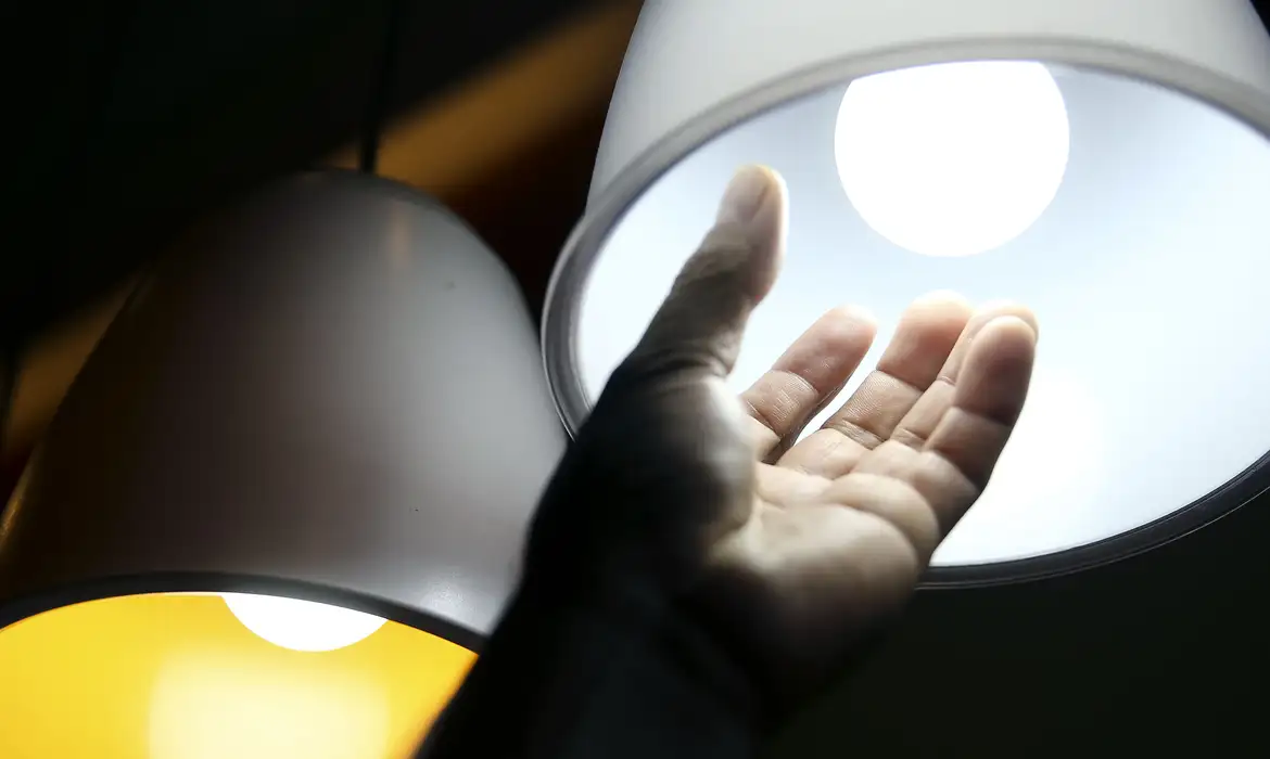 Mão alcançando uma lâmpada acesa, simbolizando a economia na conta de luz.