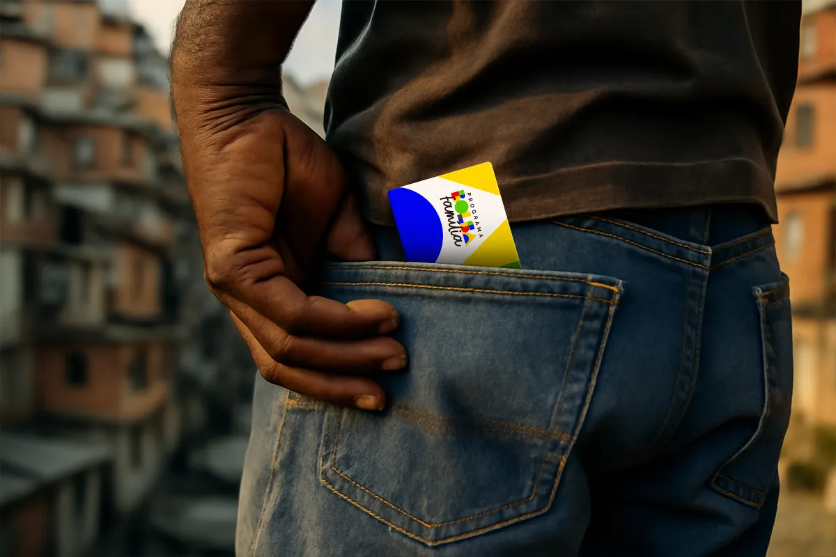 Homem colocando o cartão do Bolsa Família no bolso de trás da calça jeans, com uma comunidade ao fundo.