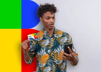 Jovem surpreso olhando para um cartão do Bolsa Família e para a tela de um celular, com um fundo vibrante em verde, azul, amarelo e vermelho.