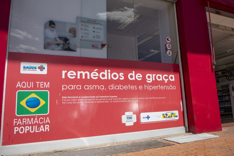Fachada de farmácia credenciada ao programa Farmácia Popular, com um banner vermelho anunciando "remédios de graça para asma, diabetes e hipertensão".
