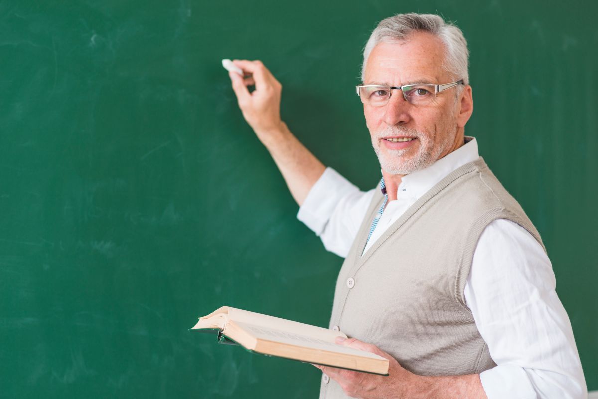 Professor sênior com óculos escrevendo no quadro-negro, representando a experiência e o planejamento da aposentadoria.