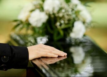 Mão repousando sobre um caixão escuro com um arranjo de flores brancas ao fundo, simbolizando o luto e a importância do auxílio funeral.