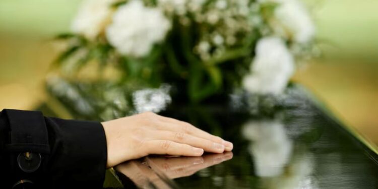 Mão repousando sobre um caixão escuro com um arranjo de flores brancas ao fundo, simbolizando o luto e a importância do auxílio funeral.