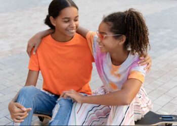 Duas garotas adolescentes, uma com camiseta laranja e outra com uma blusa tie-dye, sentadas juntas em um skate, rindo. Ambas têm o braço uma em volta da outra.