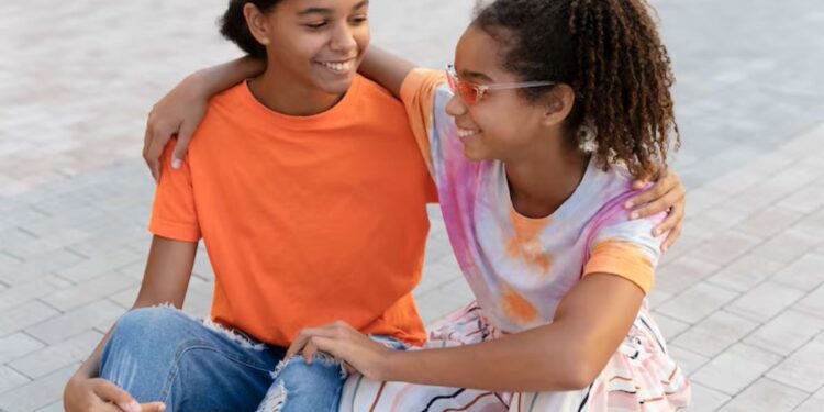 Duas garotas adolescentes, uma com camiseta laranja e outra com uma blusa tie-dye, sentadas juntas em um skate, rindo. Ambas têm o braço uma em volta da outra.