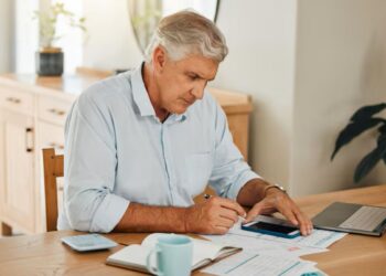 Homem de cabelos grisalhos e camisa azul claro sentado à mesa, analisando documentos e usando o celular para simular a aposentadoria no INSS.