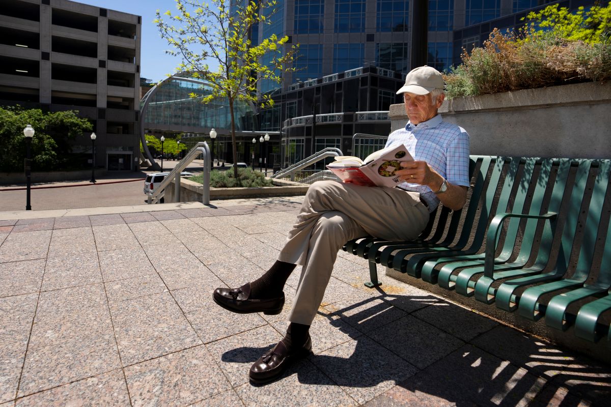 Homem idoso, sentado em um banco de praça e lendo um livro. A imagem representa o envelhecimento com qualidade de vida e a autonomia na terceira idade.